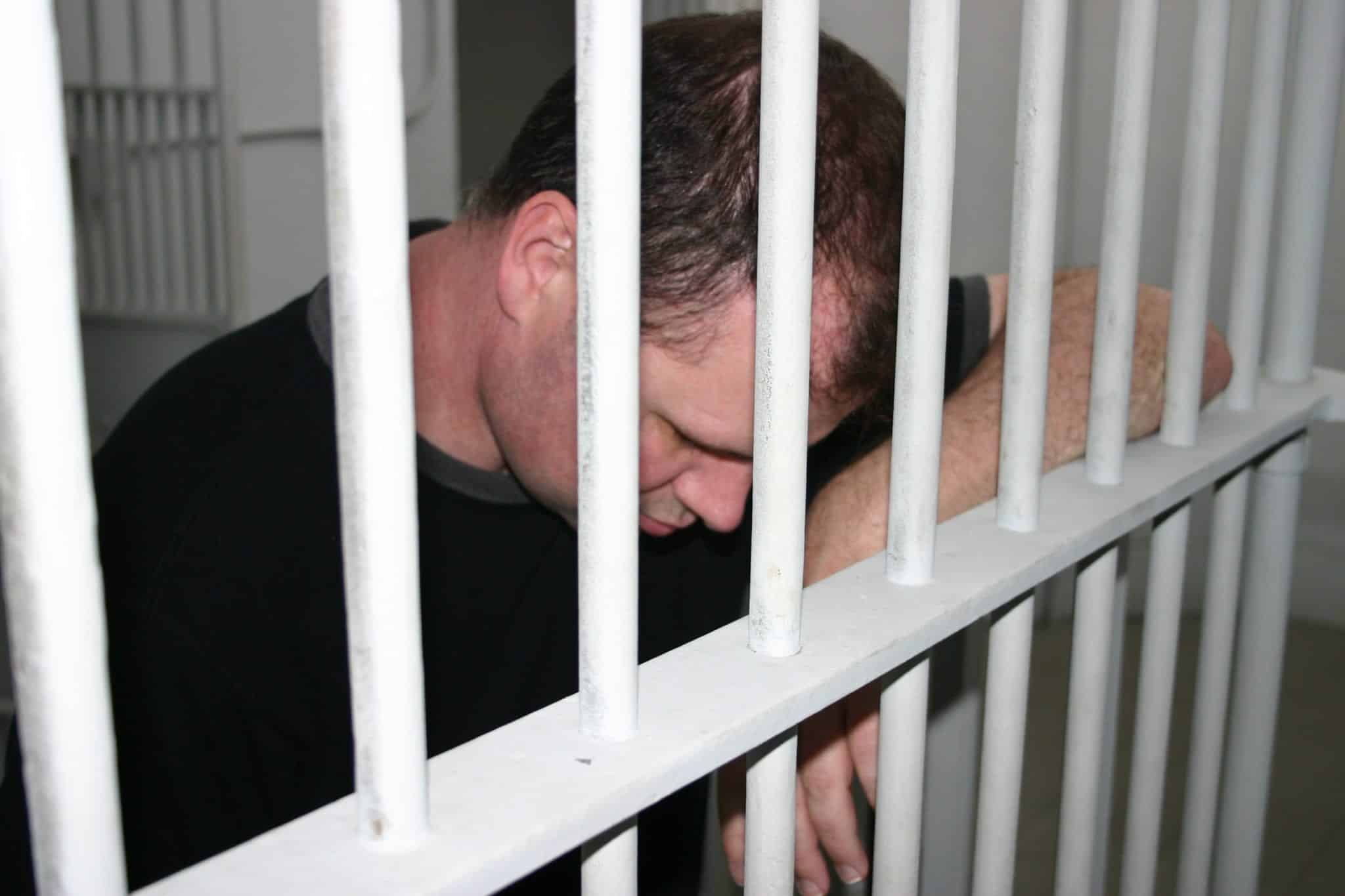 A man wearing a black t-shirt, standing in a prison cell with his hanging down and his arm propped up the white bars of the prison cell.