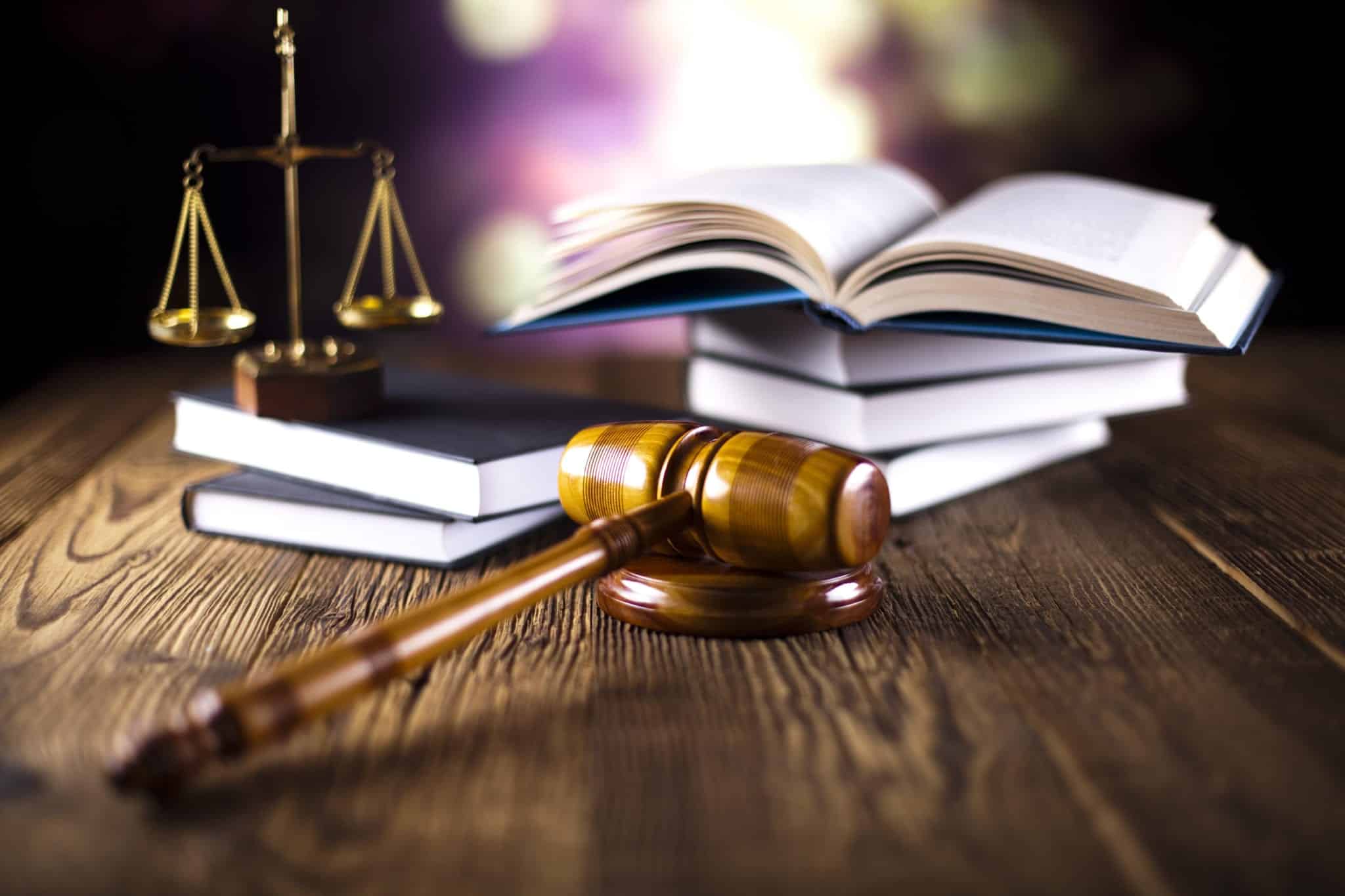 A wooden judge's gavel resting on a wooden table with stacks of books, an open book and the scales of justice blurred in the background.