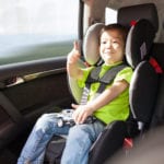 A toddler wearing a green shirt and jeans sitting in a car seat and giving a thumbs up.