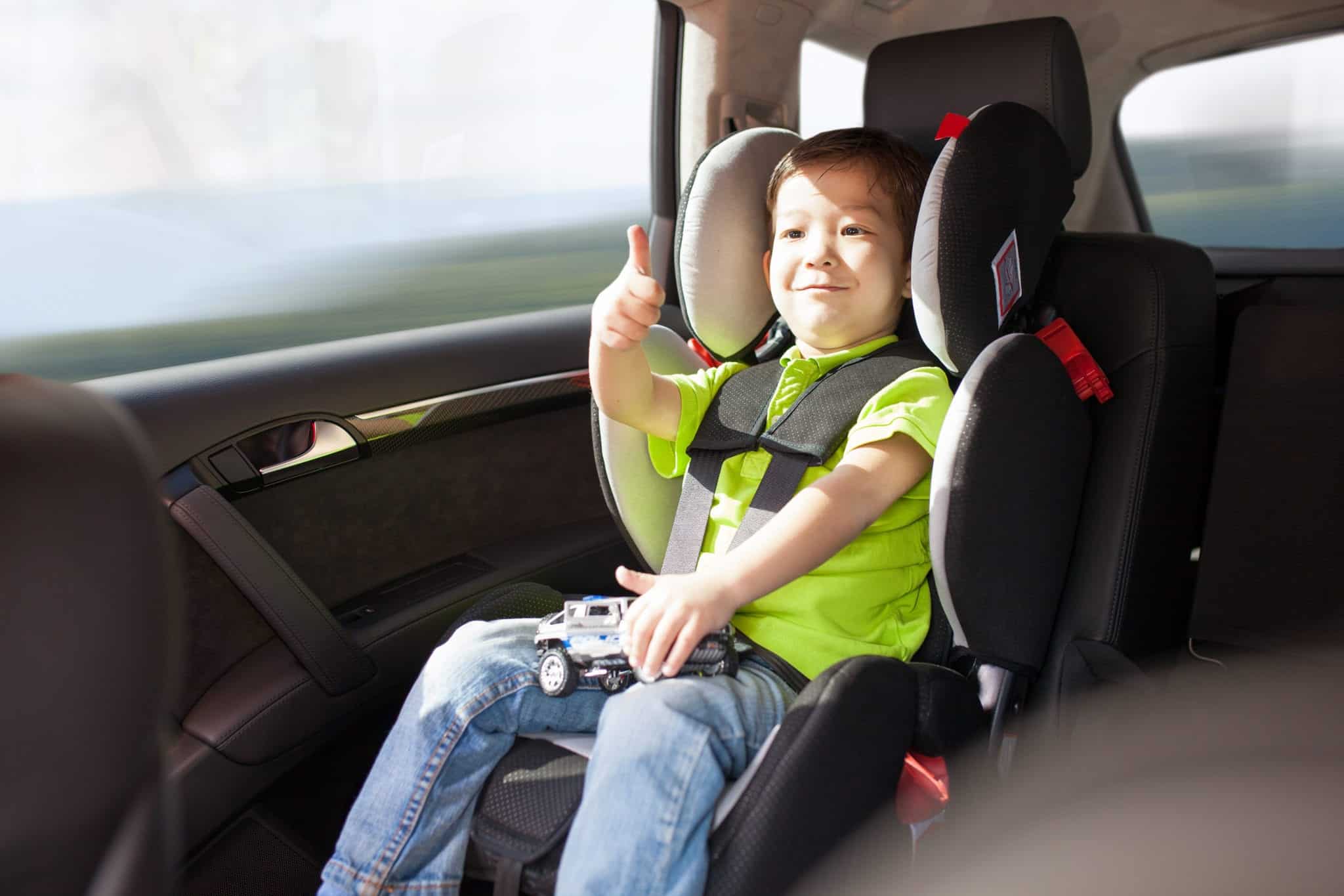 A toddler wearing a green shirt and jeans sitting in a car seat and giving a thumbs up.