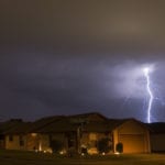 A house on a the corner of a neighborhood under a dark sky, being lit up by a streak of bright lightning.