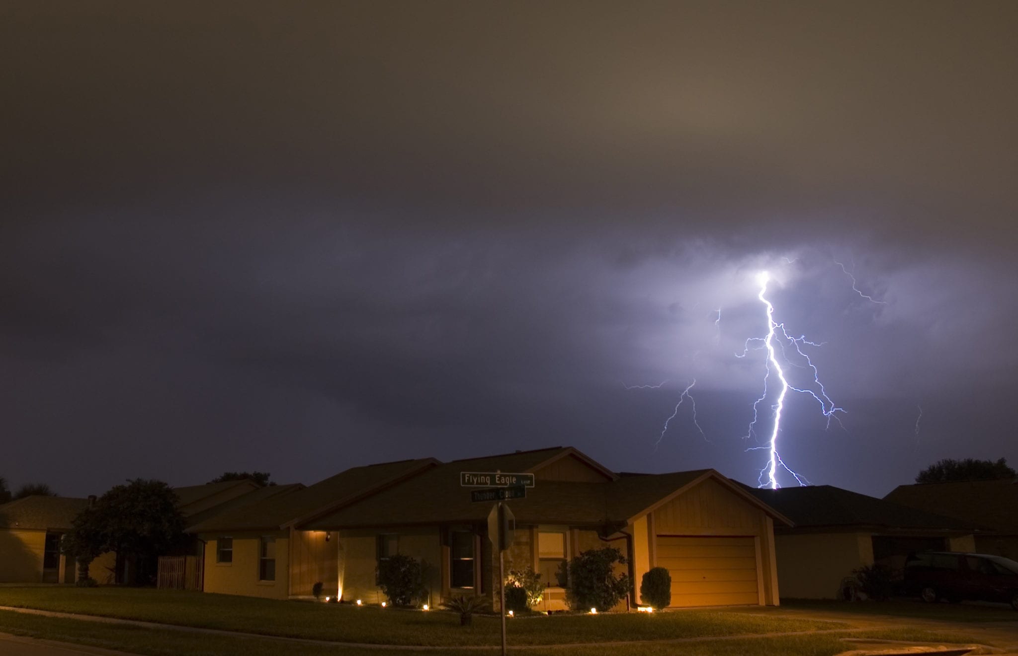 A house on a the corner of a neighborhood under a dark sky, being lit up by a streak of bright lightning.