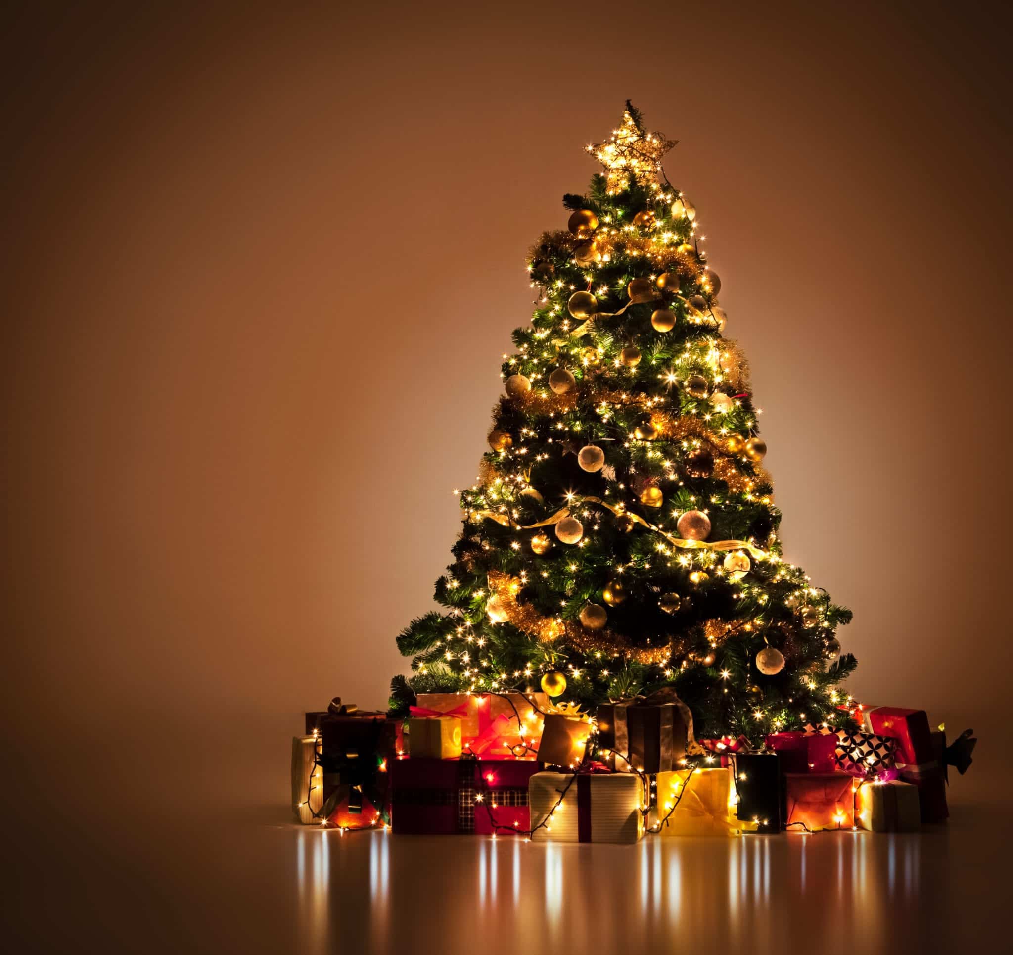 A lit up christmas tree, surrounded by gifts, in an empty beige room.