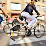 A woman in white jeans and a navy jacket riding a white bicycle through city streets with other cyclists in the blurry background.