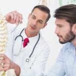 A male doctor wearing a white coat, red tie and stethoscope holding a model of the human spine and pointing to it with a ballpoint pen explaining something to the male patient on his left.