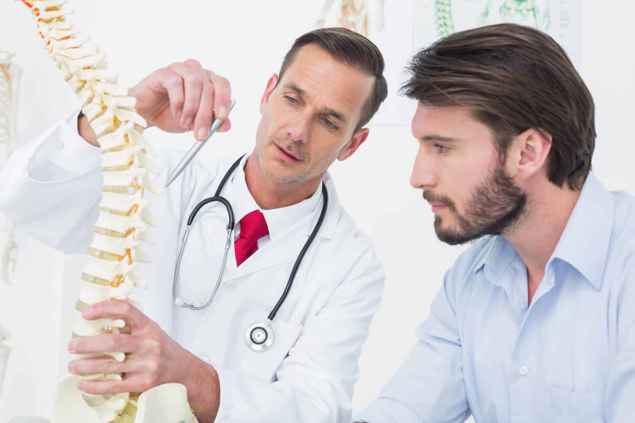 A male doctor wearing a white coat, red tie and stethoscope holding a model of the human spine and pointing to it with a ballpoint pen explaining something to the male patient on his left.