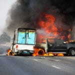 A wreck between a white delivery truck and a black pickup truck on a multiple lane road with both vehicles engulfed in flames.