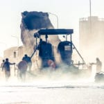 A dump truck unloading it's contents in the background with a road crew working in the middle ground and a lot of white steam coming off the road in the foreground.