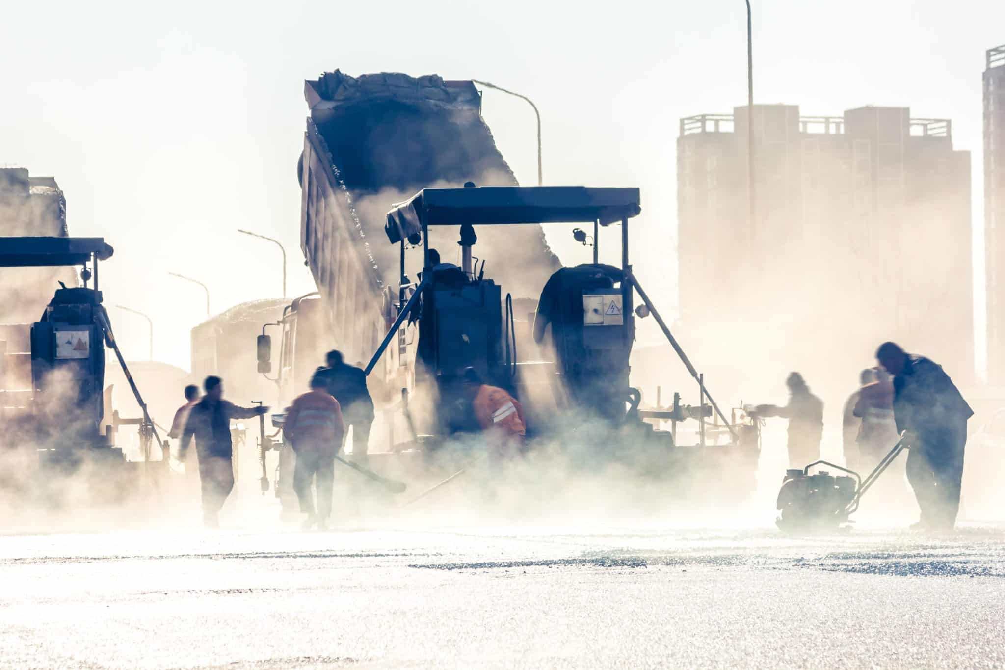 A dump truck unloading it's contents in the background with a road crew working in the middle ground and a lot of white steam coming off the road in the foreground.