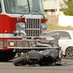 A wrecked motorcycle laying on the road in front of a red firetruck with a white car in the background.