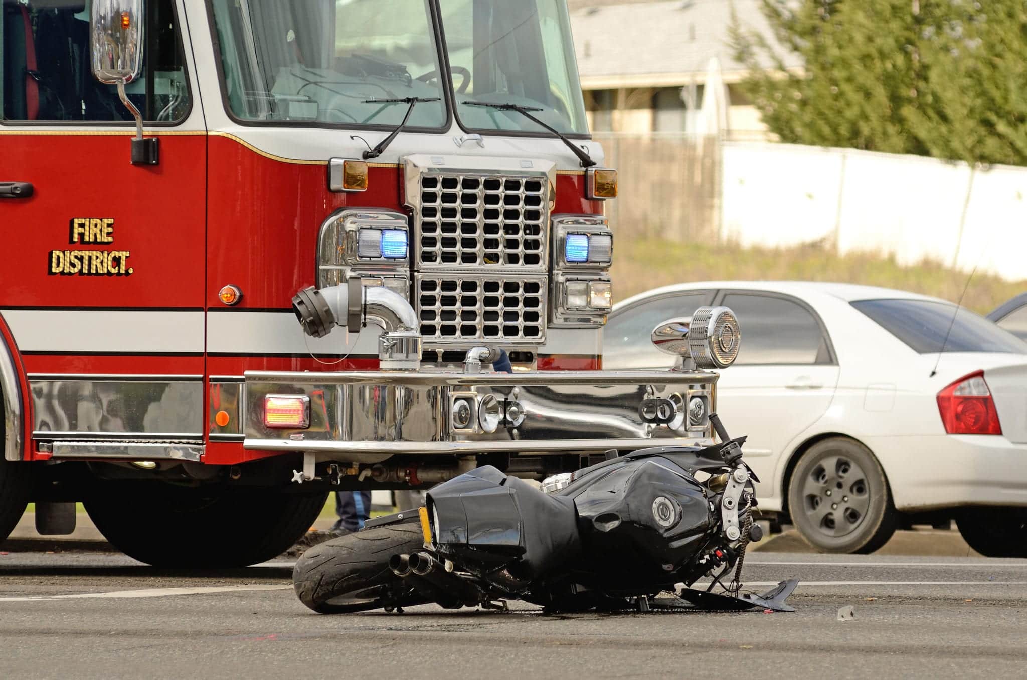 A wrecked motorcycle laying on the road in front of a red firetruck with a white car in the background.