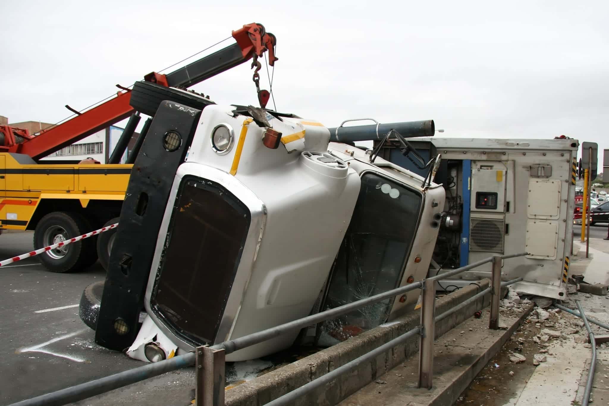 A white tractor trailer wrecked on the road and laying on it's side near a concrete curb with a metal railing, a yellow truck with a red crane extending over it appearing to be attached to the overturned truck, trying to pull it upright.