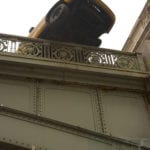 Looking up at a gray colored metal bridge and the underside of a yellow car that has driven onto the railing of the bridge and stopped before falling off the bridge completely.