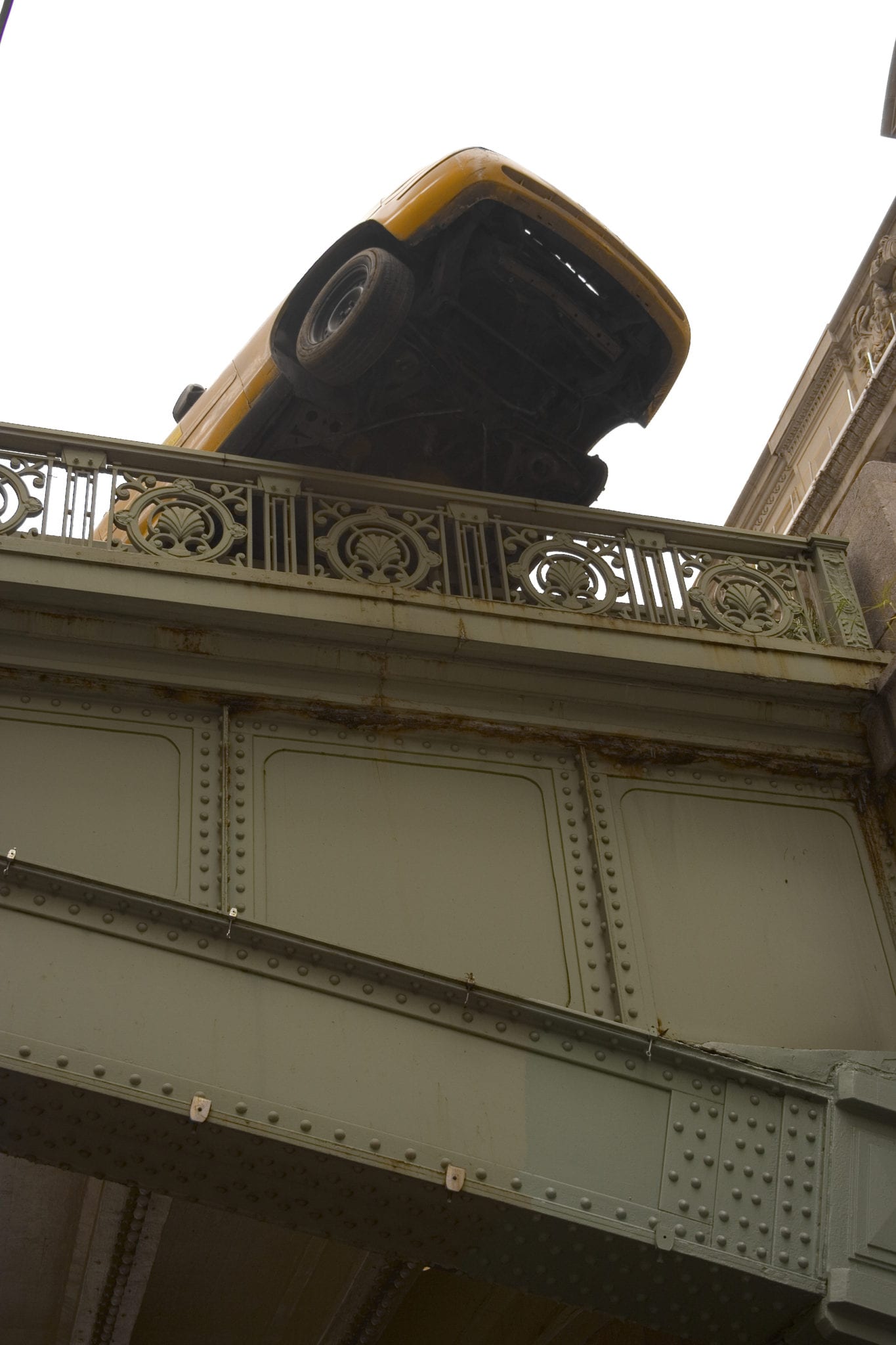 Looking up at a gray colored metal bridge and the underside of a yellow car that has driven onto the railing of the bridge and stopped before falling off the bridge completely.