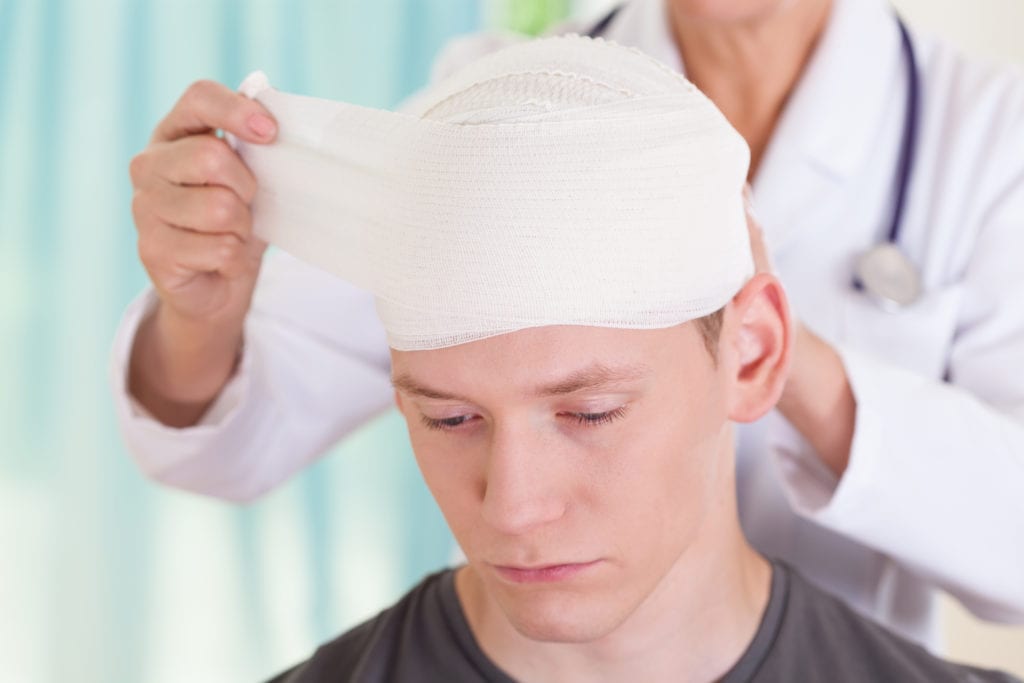 A teenage boy getting his head wrapped with a bandage by a doctor.