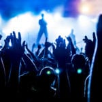 A crowd of people with their arms in the air shown in silhouette against a white foggy, blurred stage with a lead singer standing in the middle of the stage.