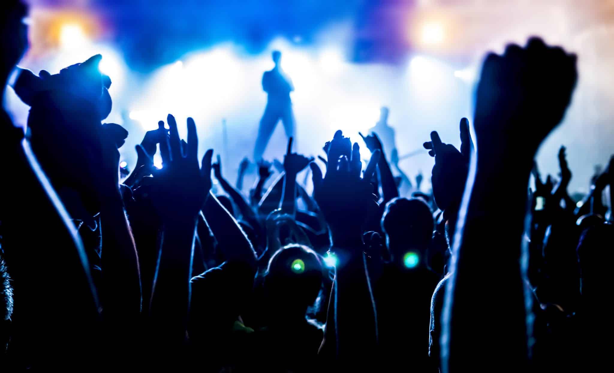 A crowd of people with their arms in the air shown in silhouette against a white foggy, blurred stage with a lead singer standing in the middle of the stage.