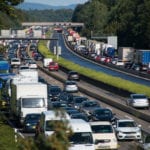 Multiple lanes of heavy traffic heading in both directions on a highway with thick green trees on each side and small mountains off in the far distance.