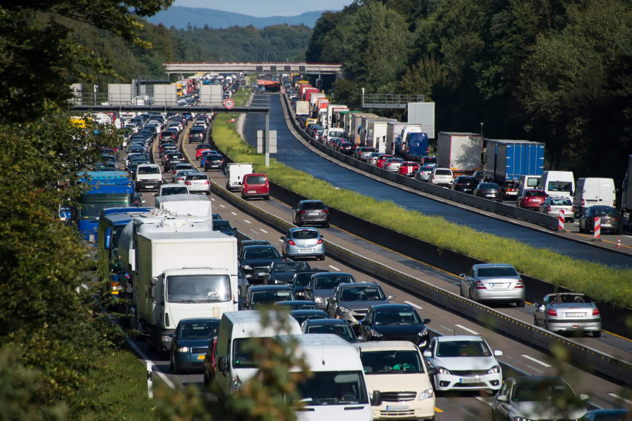 Multiple lanes of heavy traffic heading in both directions on a highway with thick green trees on each side and small mountains off in the far distance.