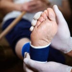 A blurry view of a woman in the background holding a wooden cane with a close-up view of her foot, wearing a blue ankle brace and beling held by a doctors white gloved hands inthe foreground.