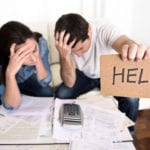 A stressed man and woman looking over a calculator and bills, with the man holding up a small piece of brown cardboard with the word HELP written on it in black marker.