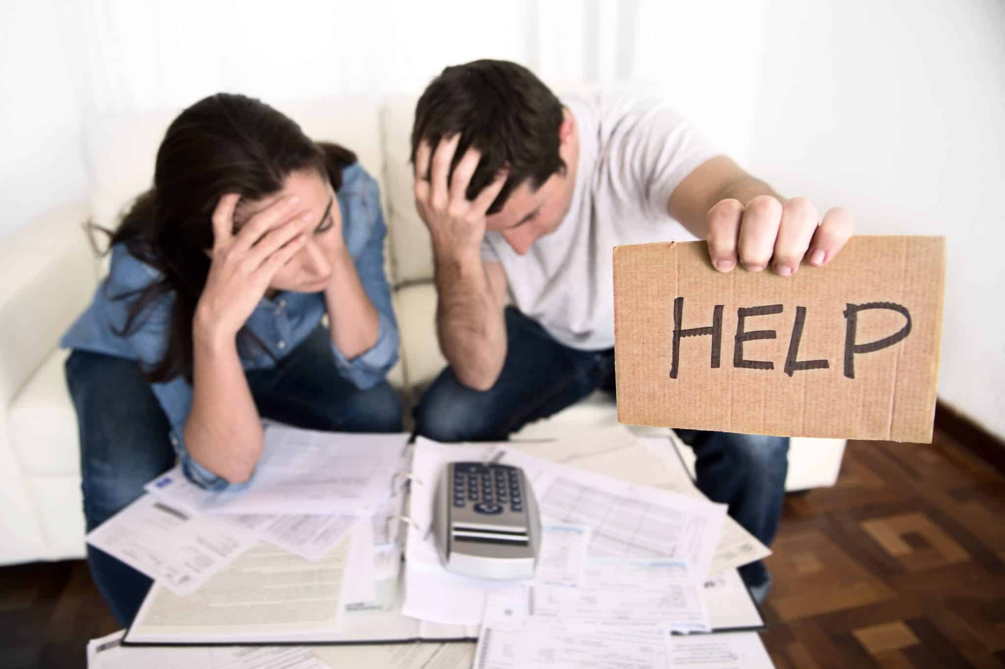 A stressed man and woman looking over a calculator and bills, with the man holding up a small piece of brown cardboard with the word HELP written on it in black marker.