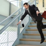 A man in a black business suit falling down stairs dropping a brown leather suitcase and a black cell phone.