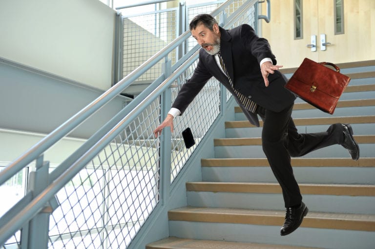 A man in a black business suit falling down stairs dropping a brown leather suitcase and a black cell phone.