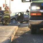 A close-up view of the right, front side of an ambulance with firefighters tending to a wreck scene in the blurry background.