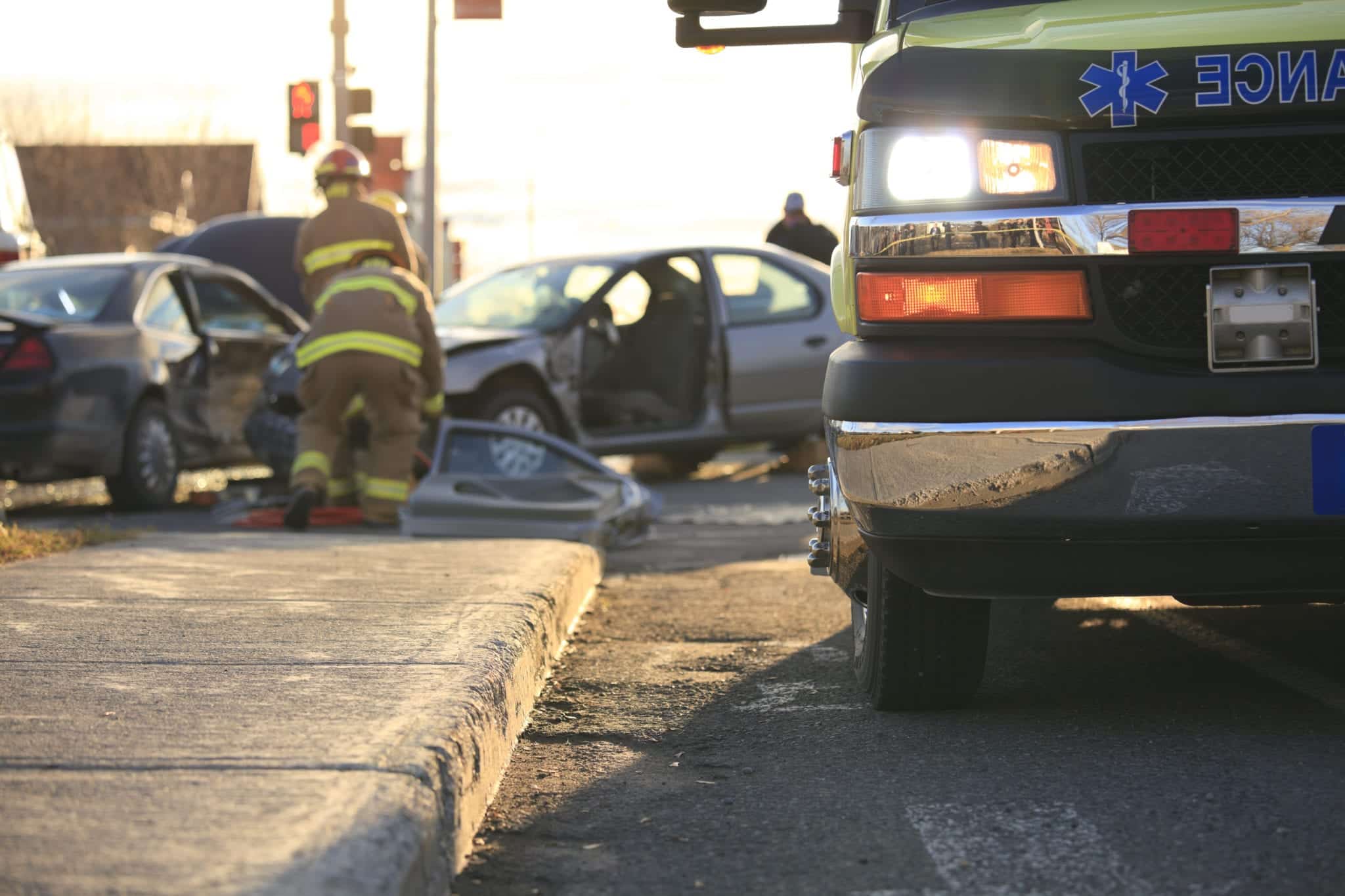 A close-up view of the right, front side of an ambulance with firefighters tending to a wreck scene in the blurry background.