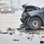 The side view of a black car with a smashed front end after a wreck, and traffic driving by in the distance.