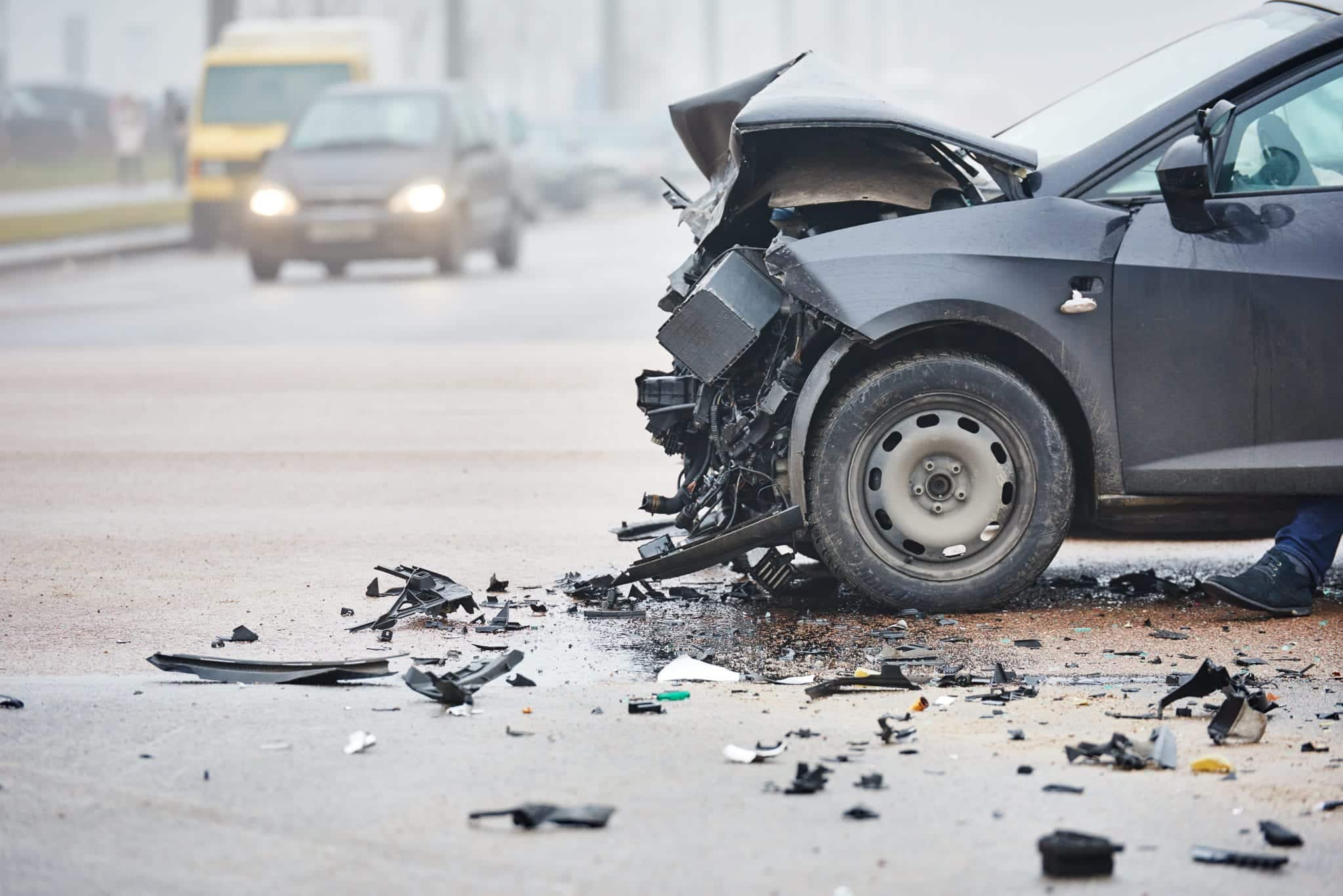 The side view of a black car with a smashed front end after a wreck, and traffic driving by in the distance.