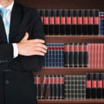 A man in a black suit, white shirt and blue tie standing, with his arms folded, in front of a shelves with several books.