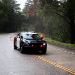 A black and white police car driving around a curve in the middle of a road surrounded by trees.