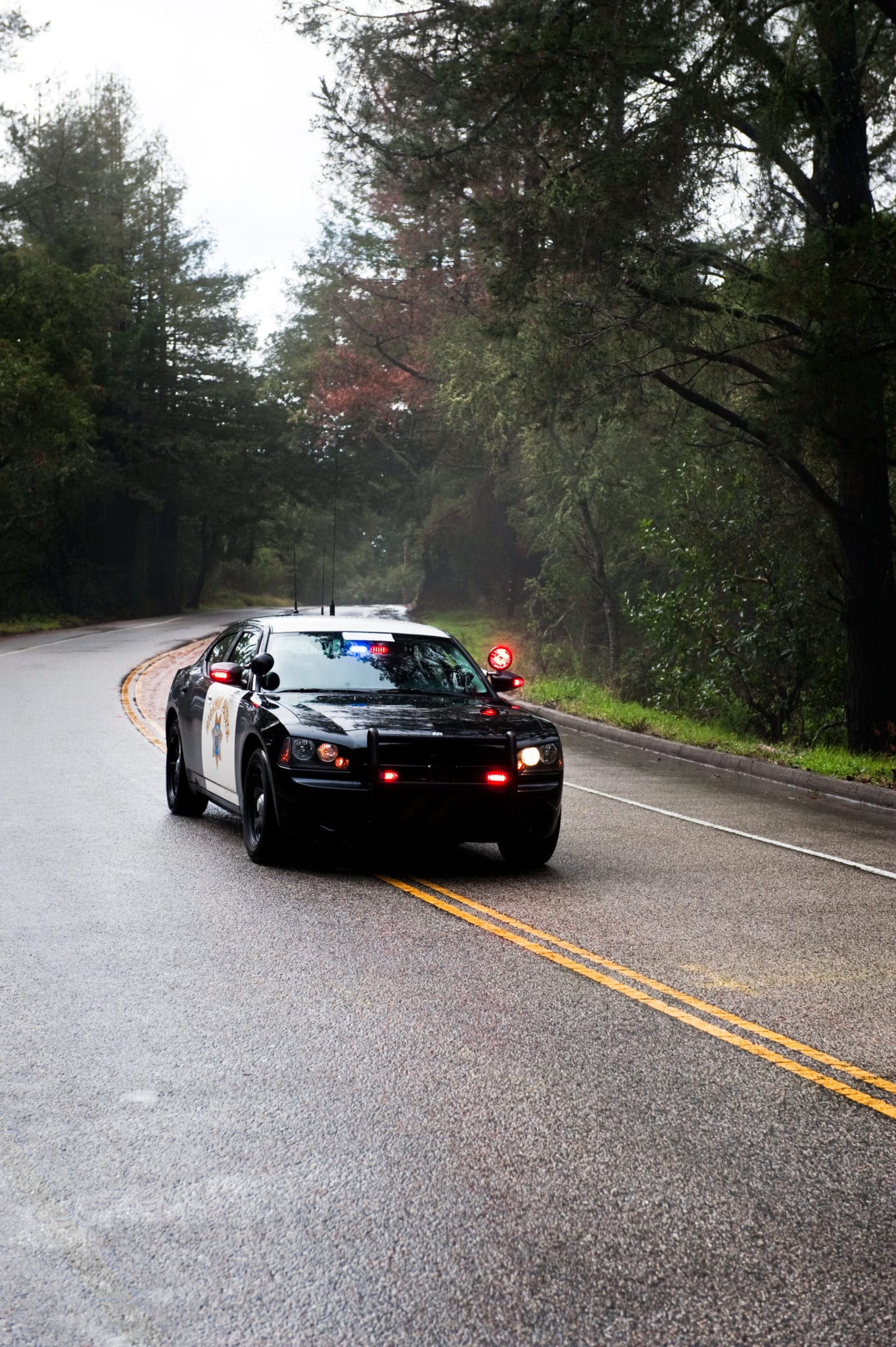 A black and white police car driving around a curve in the middle of a road surrounded by trees.