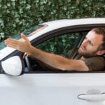 A frustrated man driving a white car, in front of a green background, with his head and arm out the window.