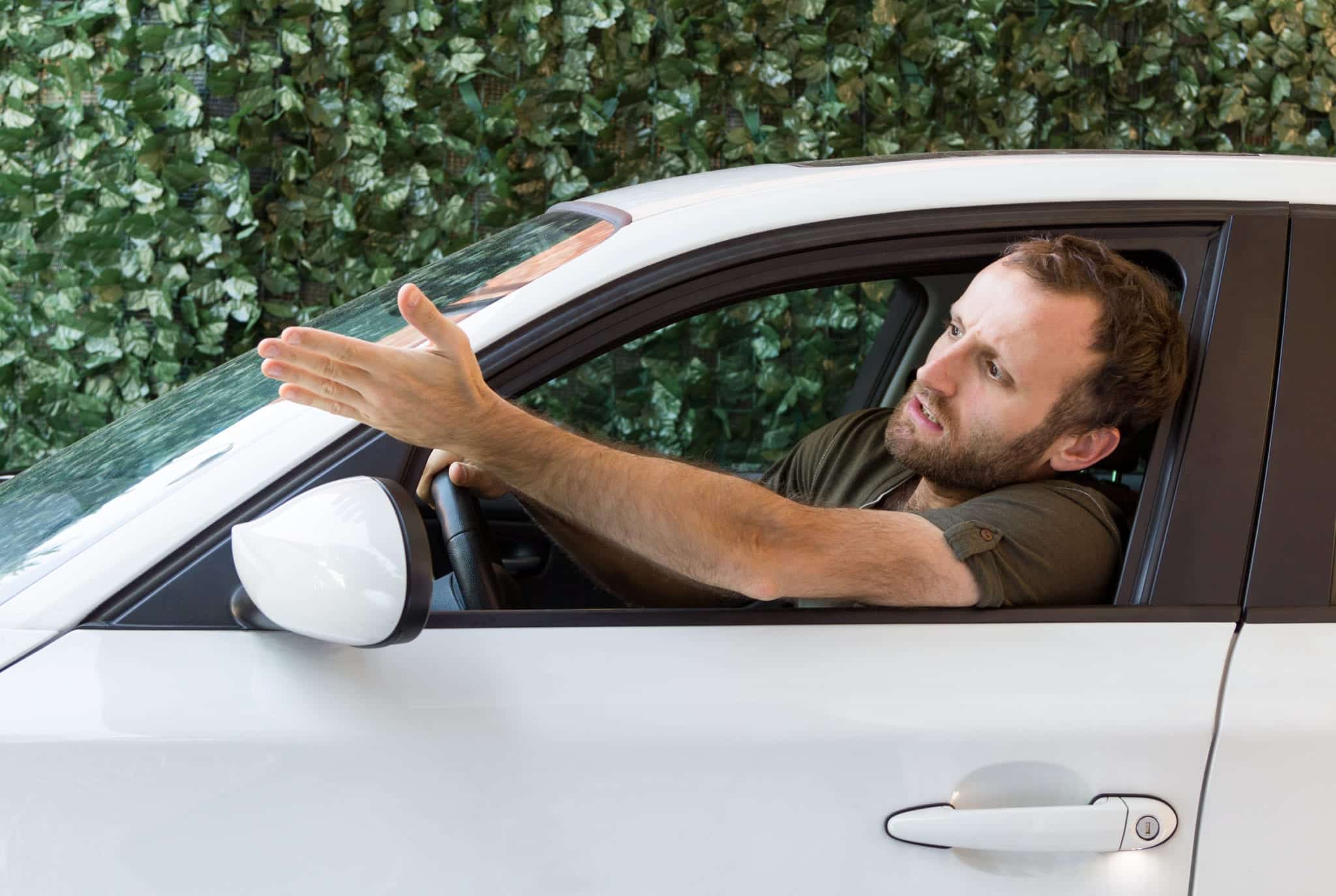 A frustrated man driving a white car, in front of a green background, with his head and arm out the window.