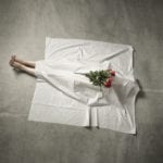 A white sheet drapped over a body laying on concrete with the lower legs exposed and a dozen red roses laying on top of the sheet.