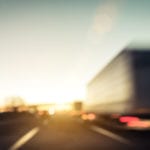 A blurry picture of a tractor trailer driving on a road surrounded by other traffic with a sunset in the distance.