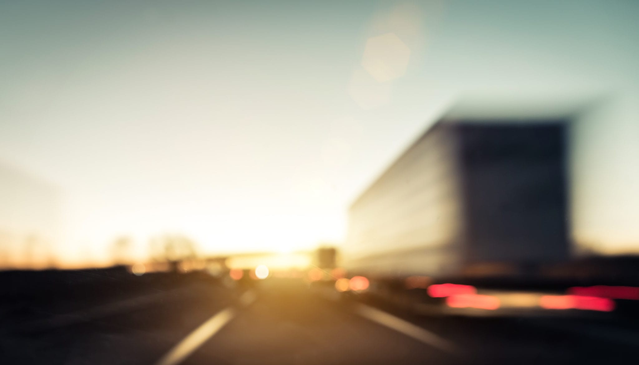 A blurry picture of a tractor trailer driving on a road surrounded by other traffic with a sunset in the distance.