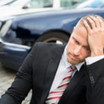 A man wearing a black suit with a red and gray stripped tie, looking disappointed with his head rested on his left hand, sitting outside on the concrete with cars in the blurred background.