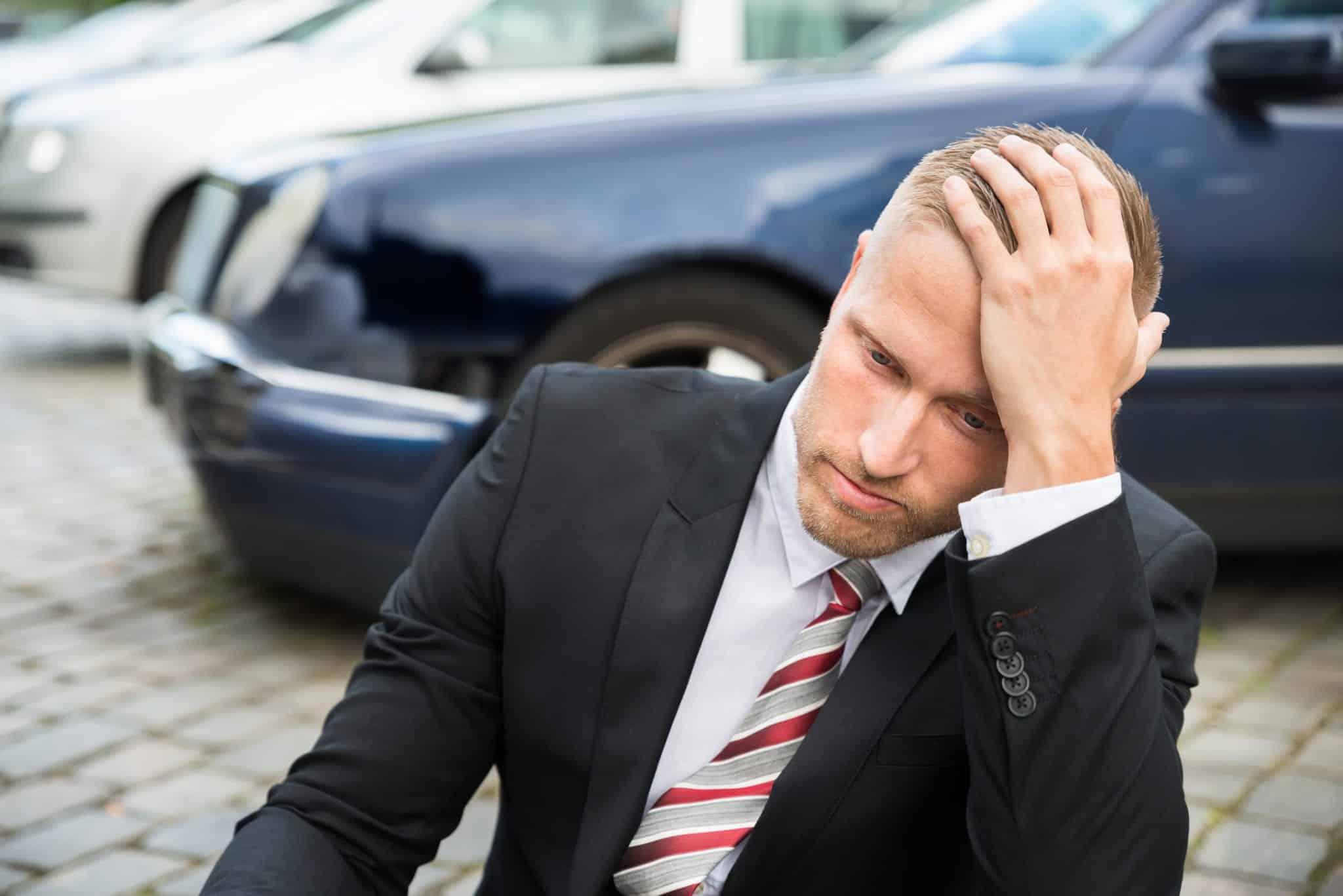 A man wearing a black suit with a red and gray stripped tie, looking disappointed with his head rested on his left hand, sitting outside on the concrete with cars in the blurred background.