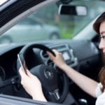 A woman texting on a cellphone with her left hand while she steers a car with her right hand.