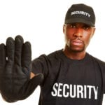 A white background with a man wearing a black t-shirt and hat with the word SECURITY written across both, a black glove on his right hand that he is holding up to indicate stop to anyone approaching.