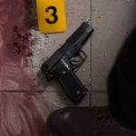 A gun lying on a tiled floor beside smear blood, broken glass, a yellow evidence tag and the foot of a man wearing dress shoes.