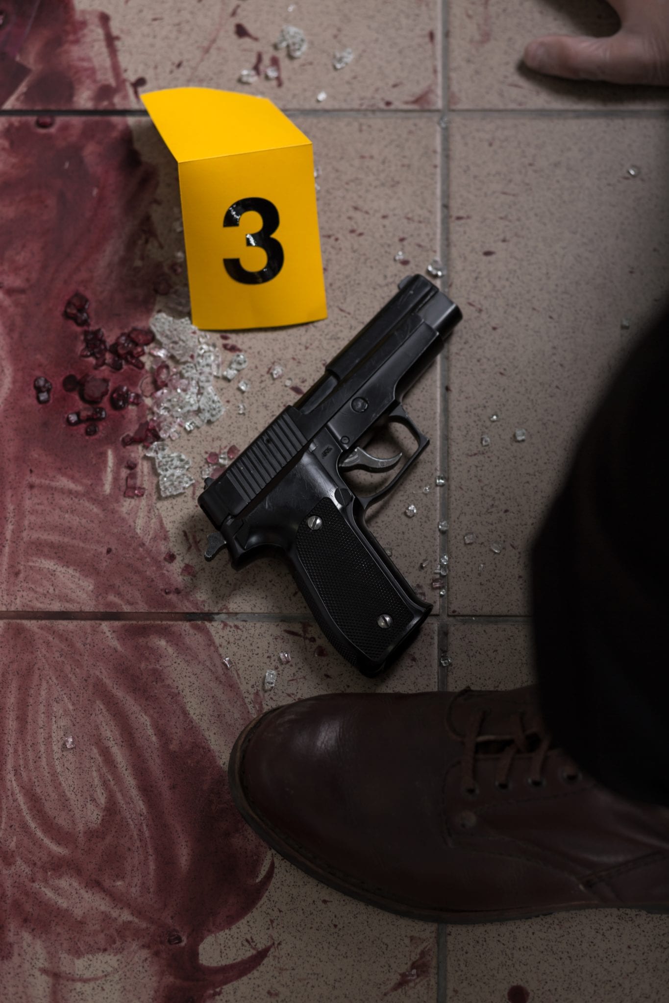 A gun lying on a tiled floor beside smear blood, broken glass, a yellow evidence tag and the foot of a man wearing dress shoes.