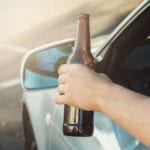 A person's arm holding a brown glass beer bottle outside the drivers side car window.