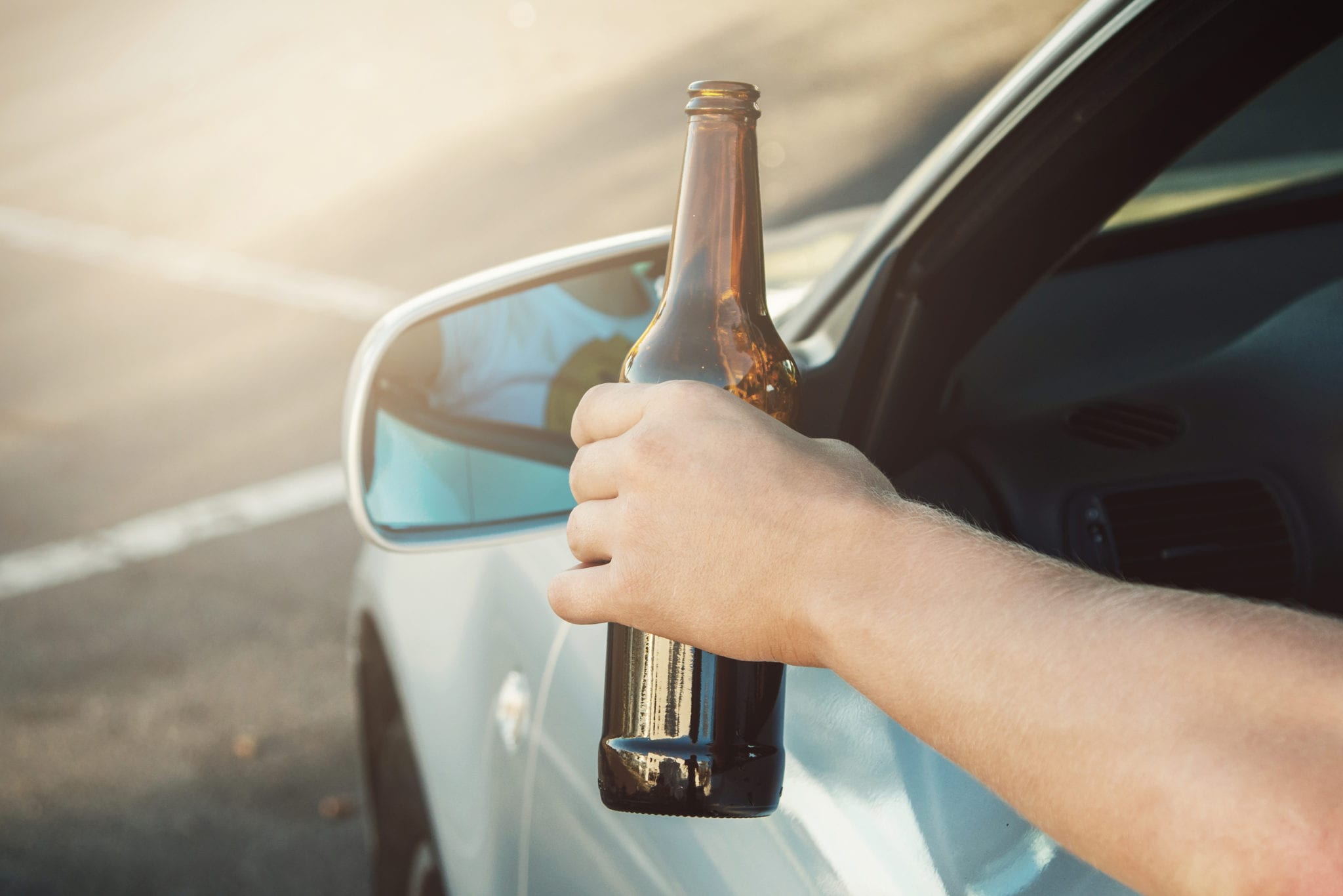 A person's arm holding a brown glass beer bottle outside the drivers side car window.