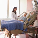 An older woman in a hospital bed with a blue blanket covering her legs, the head of the bed is at a 45 degree angle and the woman is looking at her female doctor standing beside her bed.