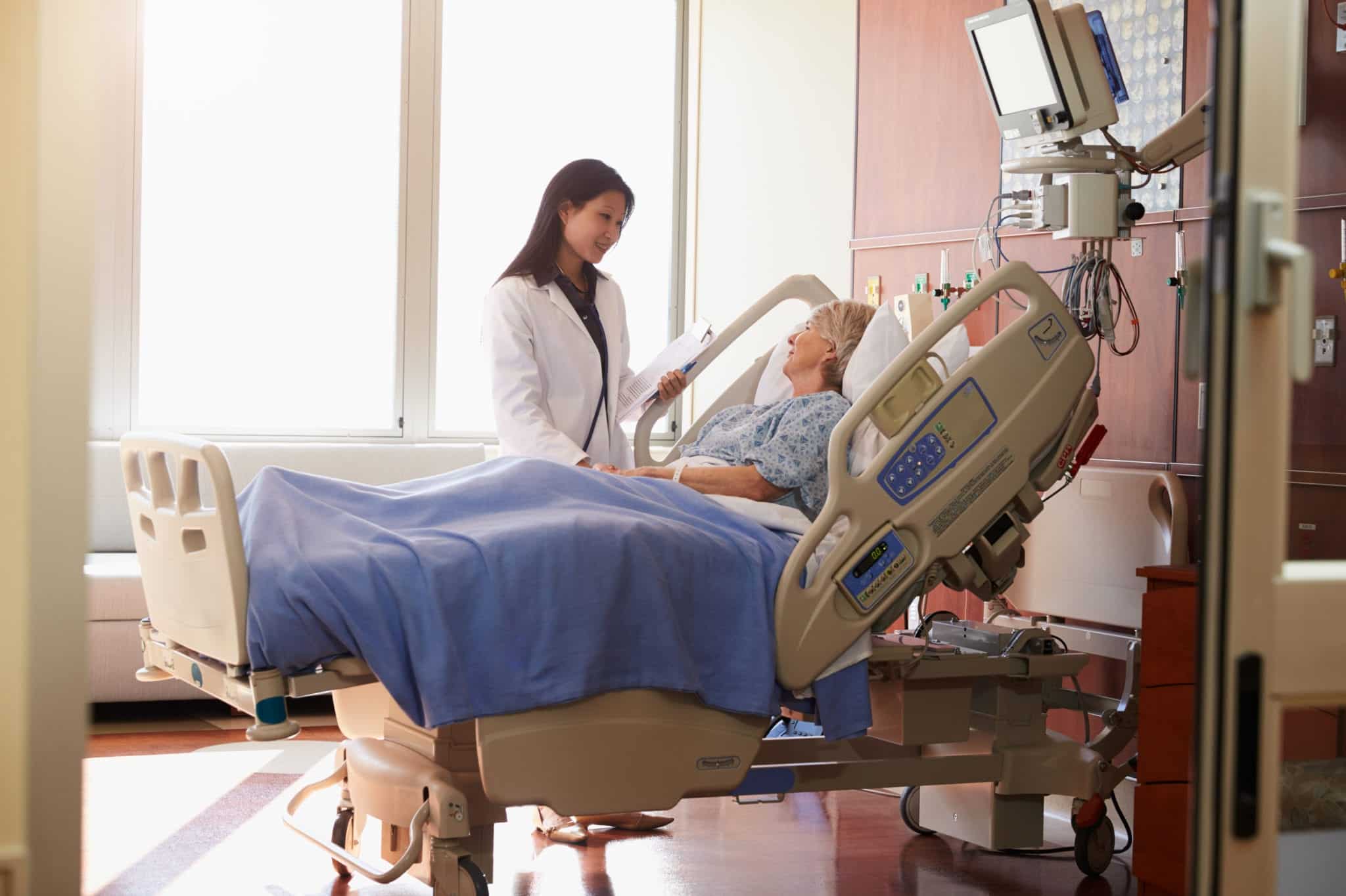 An older woman in a hospital bed with a blue blanket covering her legs, the head of the bed is at a 45 degree angle and the woman is looking at her female doctor standing beside her bed.